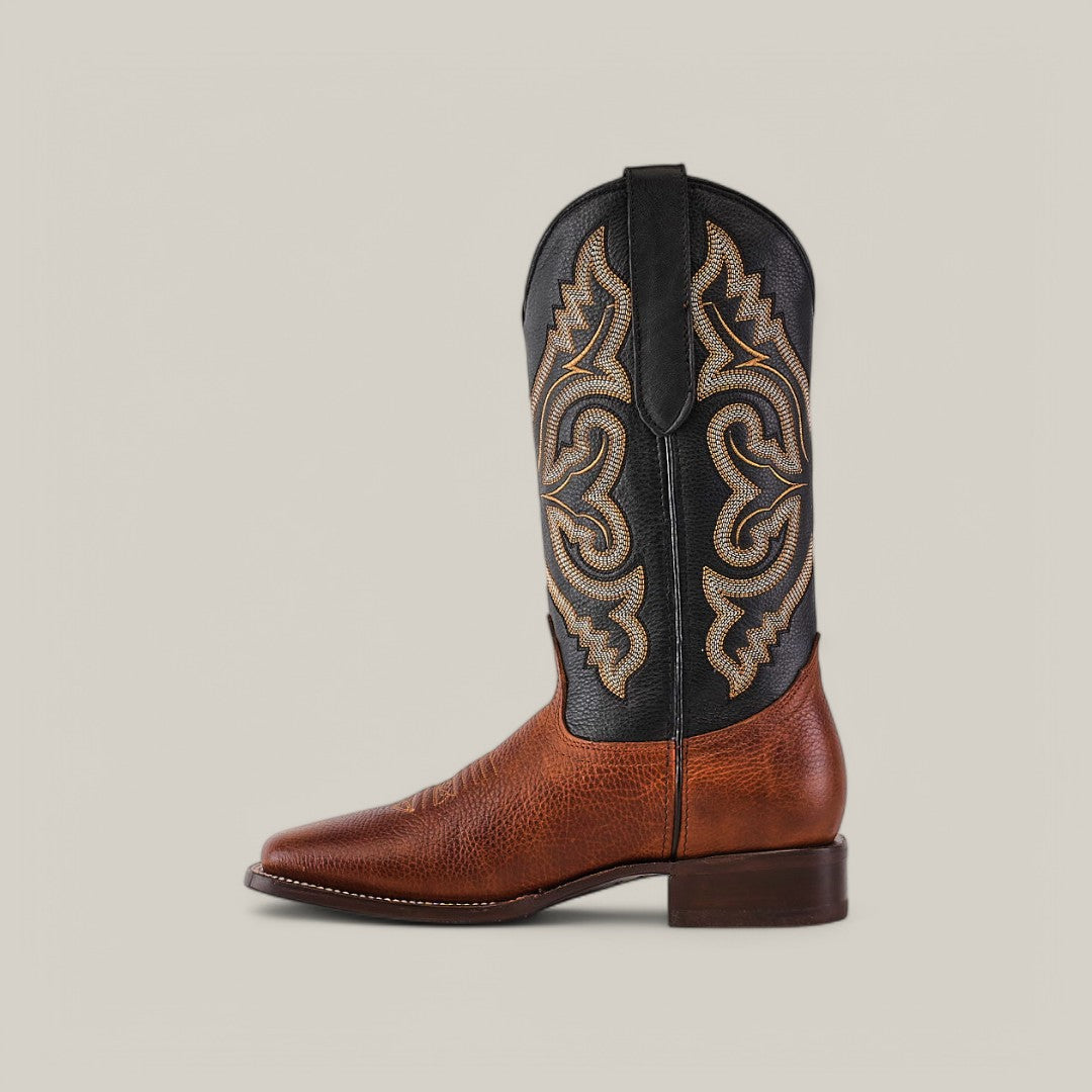 The Grisly Tan - Square Toe boot, made from premium leather, features a profile of a single cowboy boot. Its brown textured lower and decorative black upper with detailed tan stitching highlight its classic Western style against a plain gray background.