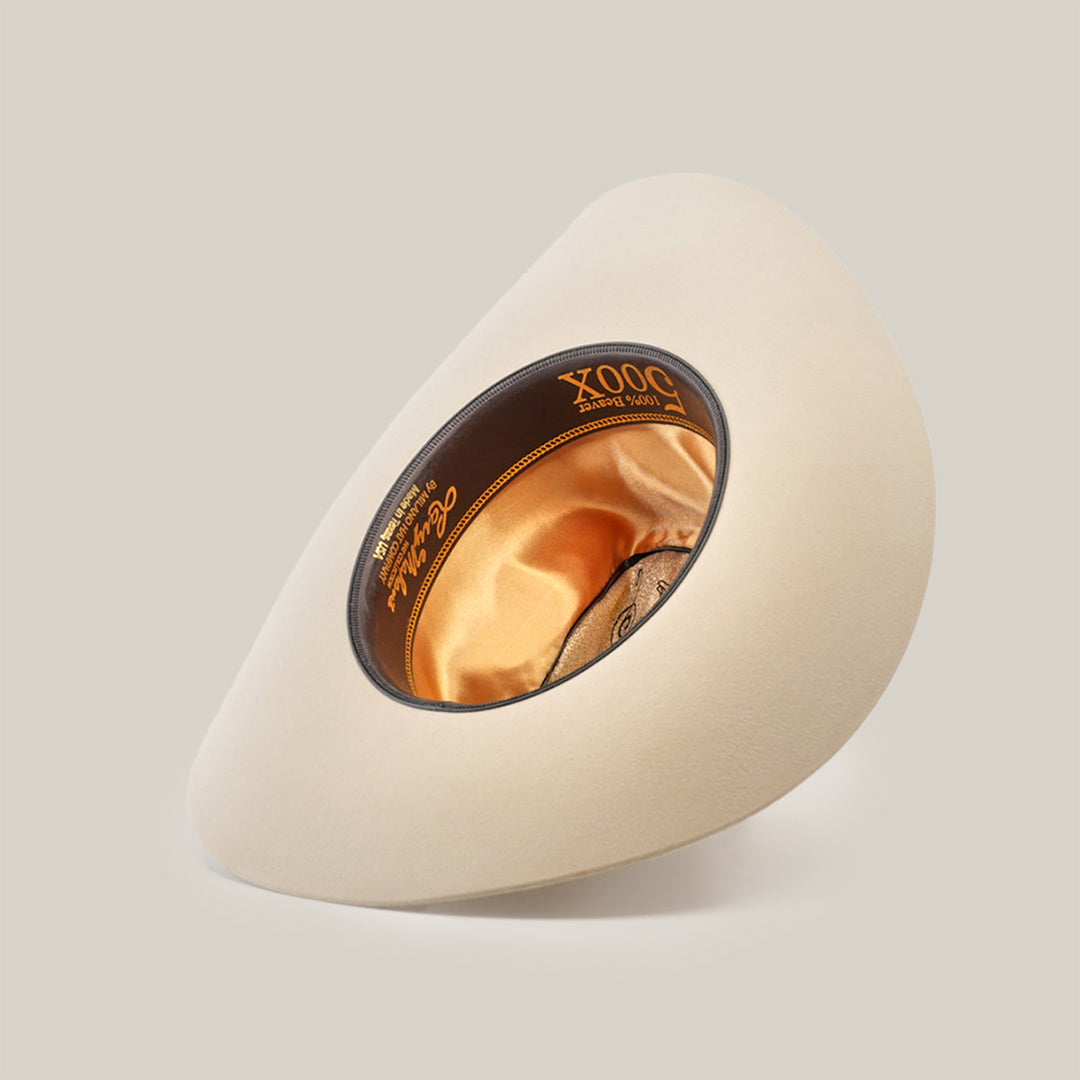 The 500X Superior Belly, a cream-colored cowboy hat made from 100% belly beaver fur, is displayed upside down to show off its satin-lined interior. A black band with gold lettering adorns the inside edge. The luxurious Western accessory contrasts against a plain white background.