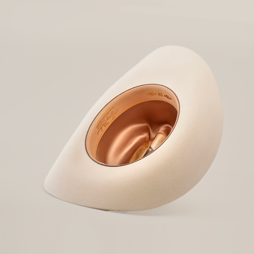 A beige felt hat, the 10X Jerarca Belly, is displayed against a white background with a tilted brim. This western headwear features shiny gold lining and an inner label, placed on its side to highlight Larry Mahans timeless style.