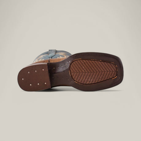 The image shows a single Sierra Cinnamon - Square Toe boot on its side, featuring a textured brown grip sole. This Western-style boot boasts intricate decorative stitching, showcasing premium leather craftsmanship against a neutral beige backdrop.