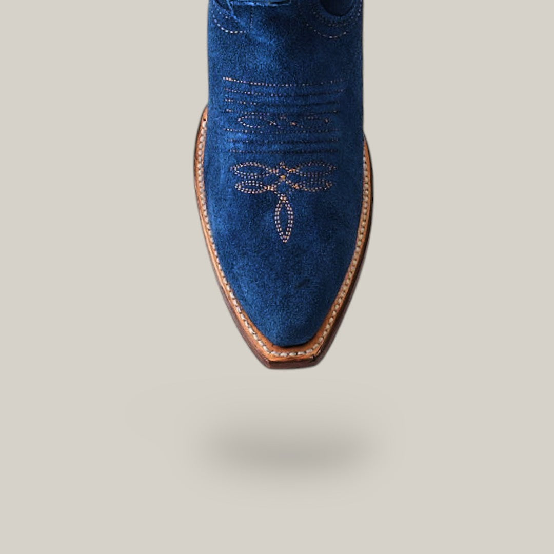 Close-up of the Miranda Suede Blue Jeans boot with snip toe and dragonfly stitching in blue suede leather. It features a leather sole and is set against a plain, light gray background.