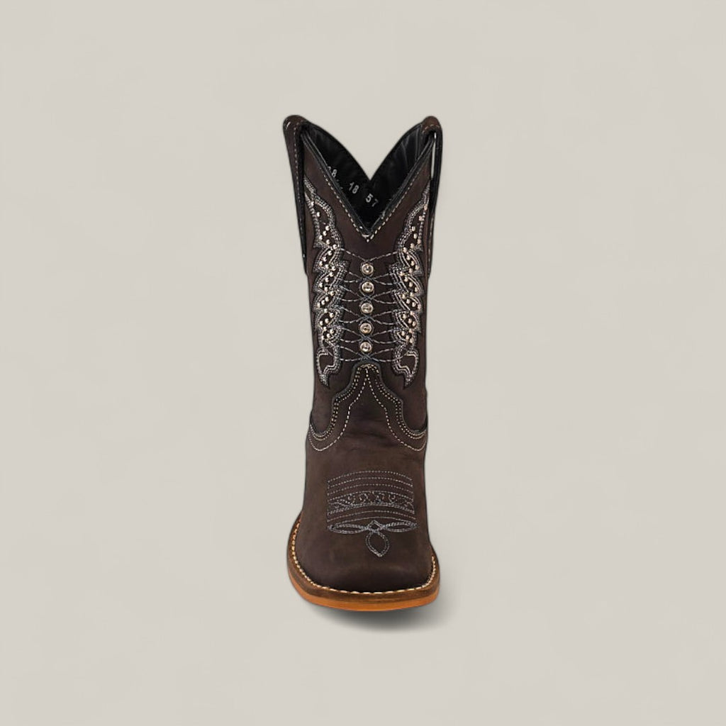 The Orion Suede Oxford - Square Toe Kids Boot is shown from the front in brown suede, featuring intricate white and silver stitching with decorative patterns, set against a plain light gray background.