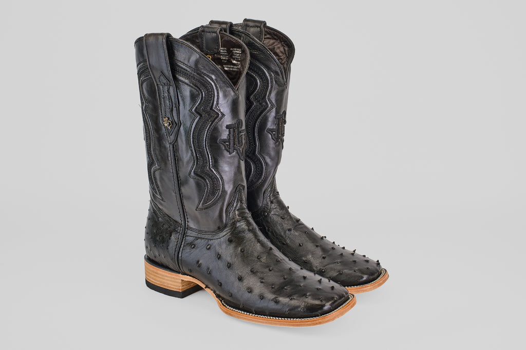 The Austin - Black - Square Toe boots feature decorative stitching, square toes, and light brown stacked heels, crafted in black avestruz leather and displayed against a plain light gray background.