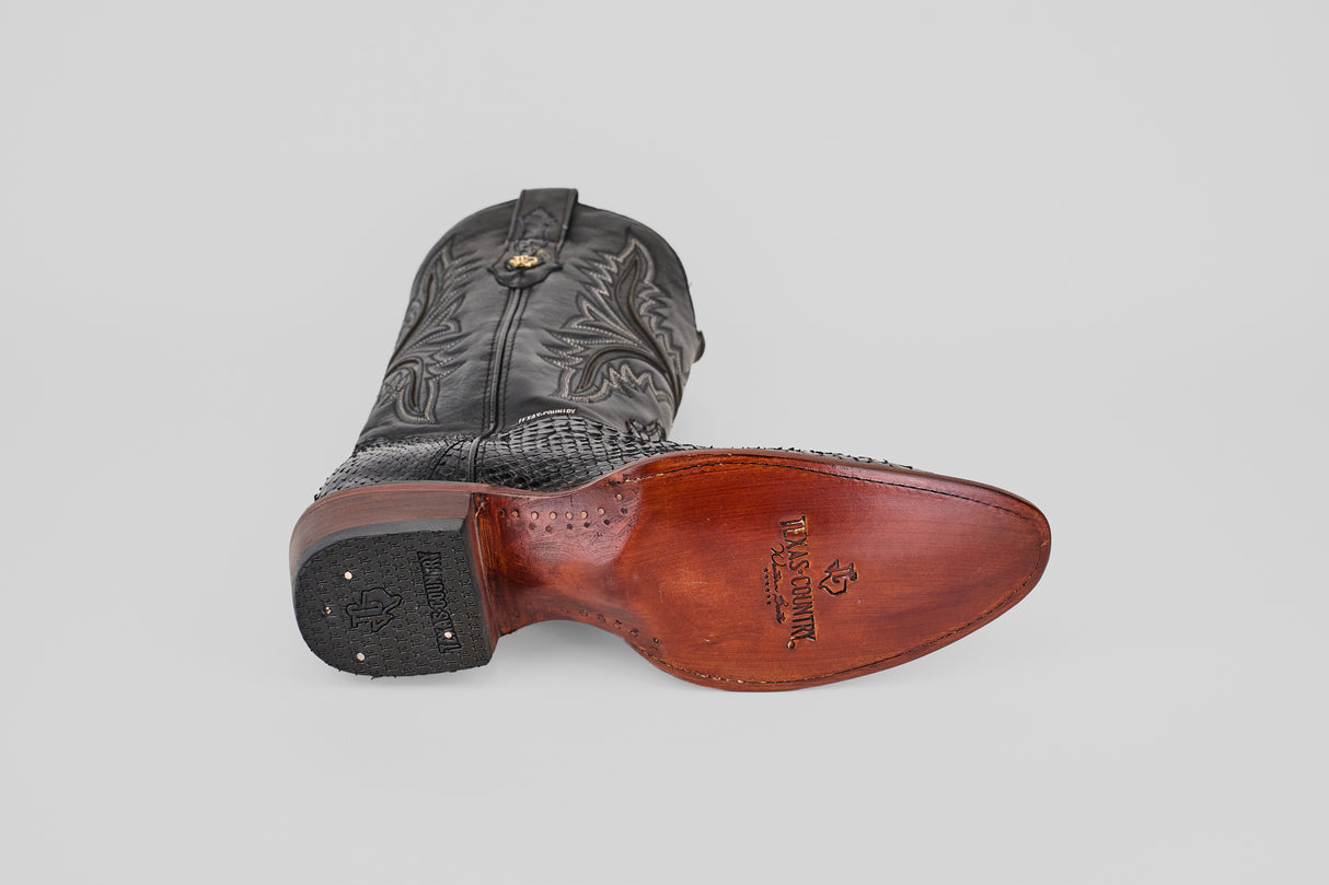 A single Tanner - Black - Round Toe cowboy boot rests on its side, highlighting detailed stitching on the upper and a smooth brown Python Negro Oval leather sole with the Black Star by Justin logo.