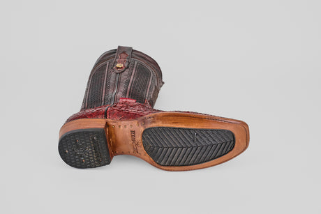 A single Tanner – Ruby – Narrow Square Toe western boot rests on its side, displaying a worn red and brown leather upper and a partially rubber textured sole, set against a plain gray background.