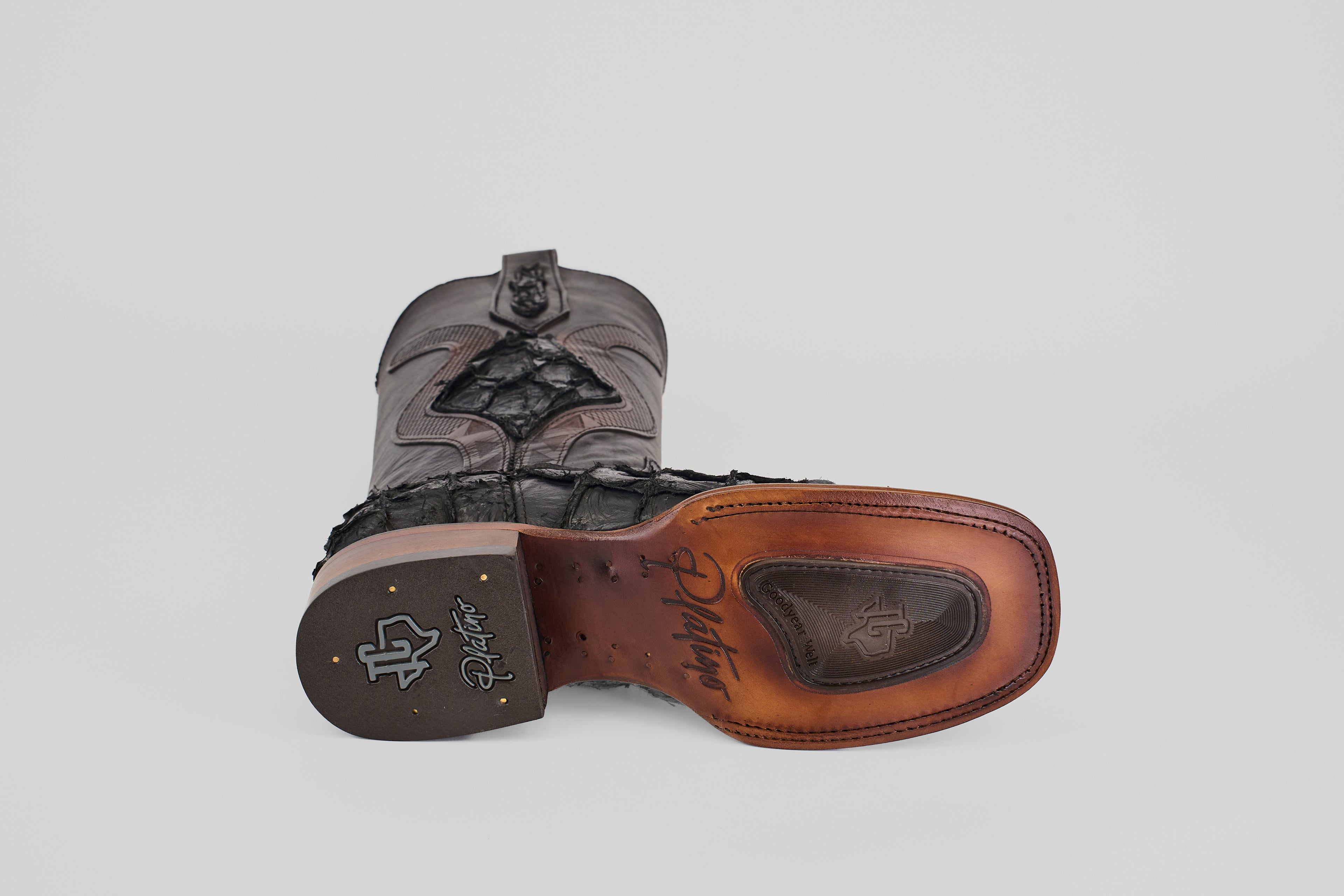 The Conor Platinum - Matte Black - Square Toe (Diamond) western boot is shown on its side, highlighting the brown leather sole with branded logos and stitching, plus a dark gray and black textured patterned upper.