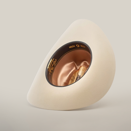 The 6X Real Belly is a beige, wide-brimmed fur felt hat, displayed upside down to reveal its satin-lined interior. The inside band features gold lettering and the number 9, all elegantly set against a light, plain background.