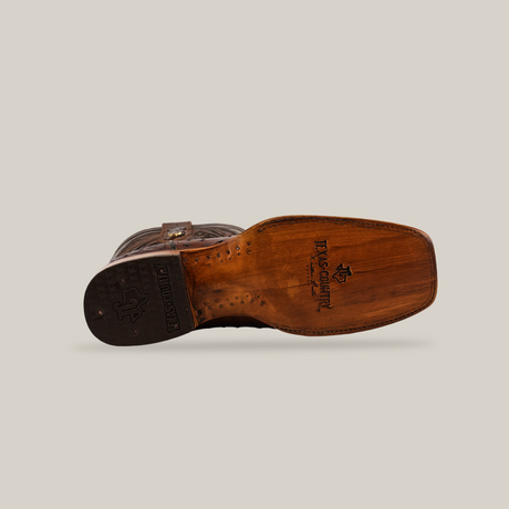The Exotic Ostrich - Brown - Rodeo Toe boot displays a wooden sole with engraved logos and signature details. Its superior craftsmanship shines through as it rests on its side against a white background, highlighting the artistry that complements its ostrich leather upper.