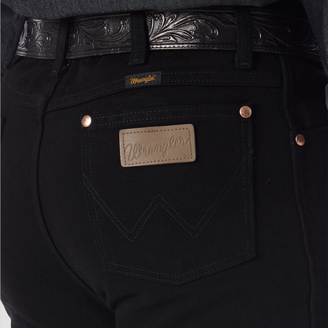 Close-up of a person wearing Wrangler Cowboy Cut Slim Fit Jean - 0936WBK with visible stitching and a branded leather tag, paired with a dark, intricately patterned leather belt.